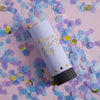 A white tube of hand twist confetti cannons with gold accents lying on a pink surface surrounded by purple and blue confetti.