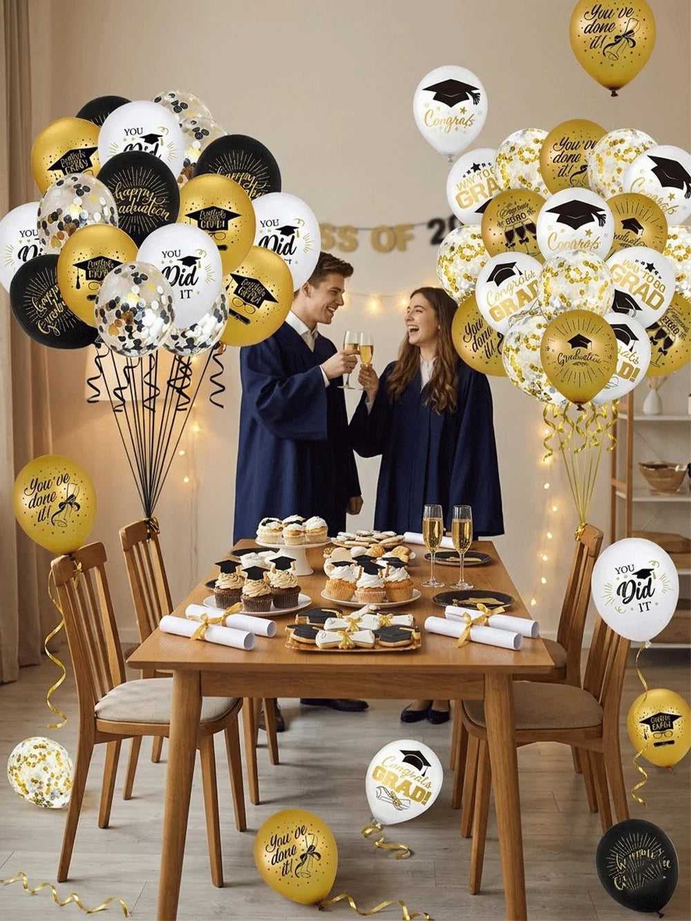 A table decorated with gold and white graduation balloons featuring caps and stars, surrounded by people.