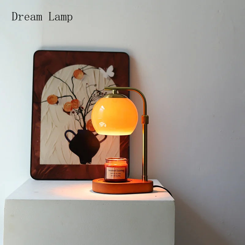 A golden aromatherapy wax melting lamp with a yellow glow sits on a white surface in front of a framed artwork.