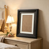 A black photo frame with an antique gold trim rests on a wooden nightstand next to a lamp.