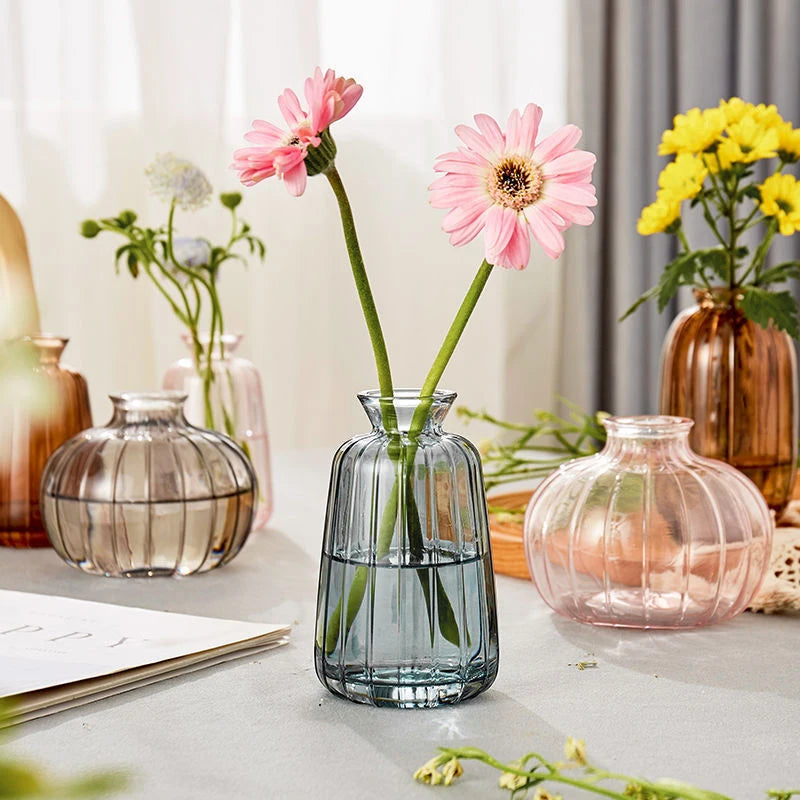 A modern clear glass vase filled with green stems and pink flowers sits on a light surface.