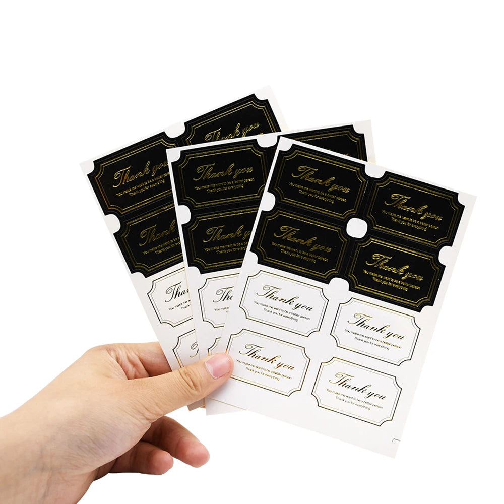 A hand holds several black thank you seal stickers for crafting and packaging.