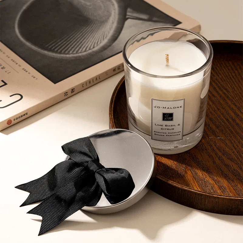 A clear glass candle cup with a white candle is placed on a wooden tray next to a black ribbon.