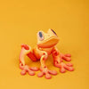 A small, orange tree frog ornament with large eyes is displayed against a plain yellow background.