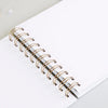 A white spiral-bound notebook with gold-colored rings lies on a light gray surface.