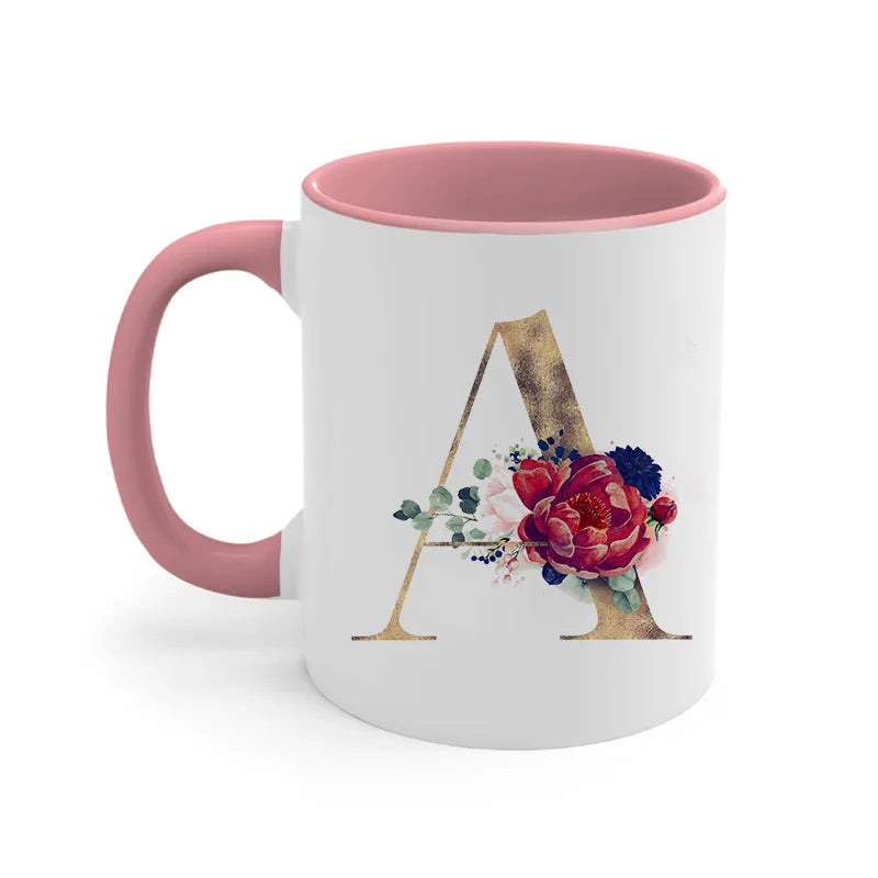 A pink ceramic coffee mug featuring a gold letter initial design surrounded by floral accents.