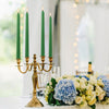 A brass candelabra holds three tall green stick candles on a white table with a bouquet of blue and cream flowers.