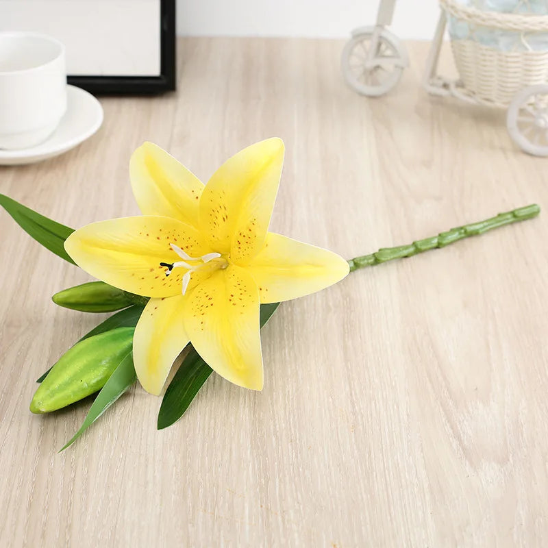 A yellow silk lily flower stem with three blooms resting on a light wooden surface.