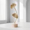 A gold-colored iron ginkgo leaf ornament set with a slender stem mounted on a light wood base.
