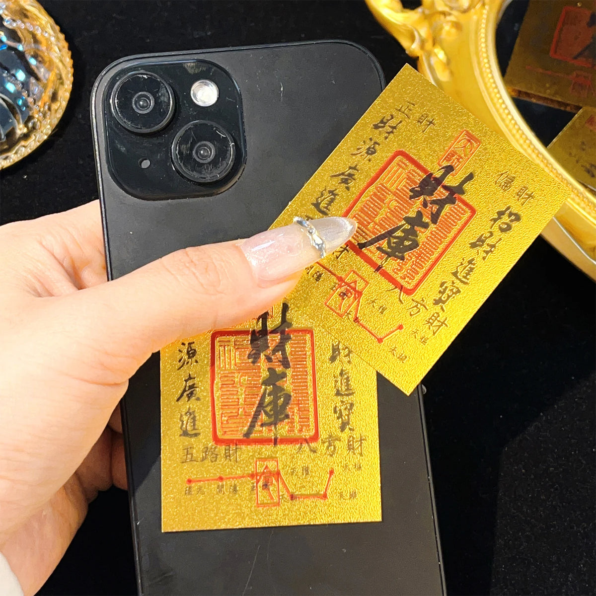A hand holds a gold wealth amulet phone sticker next to a golden object.