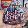 A hand holds a colorful wooden fridge magnet featuring the text Kiala Lumpur and city skyline silhouettes.