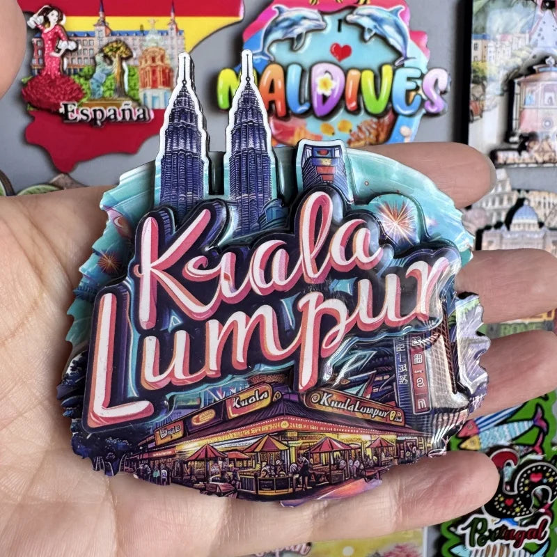 A hand holds a colorful wooden fridge magnet featuring the text Kiala Lumpur and city skyline silhouettes.