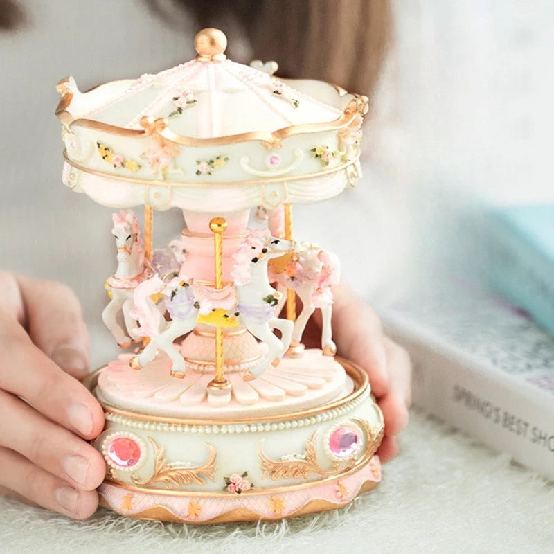 A delicate pink and white resin carousel music box with horses and floral accents is held in a person's hand.
