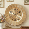 A round gold-toned wall clock with a sunburst design and Roman numerals mounted on a light wooden surface.