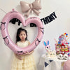A young girl holds a large pink heart-shaped balloon decoration with black ribbons against a white wall.