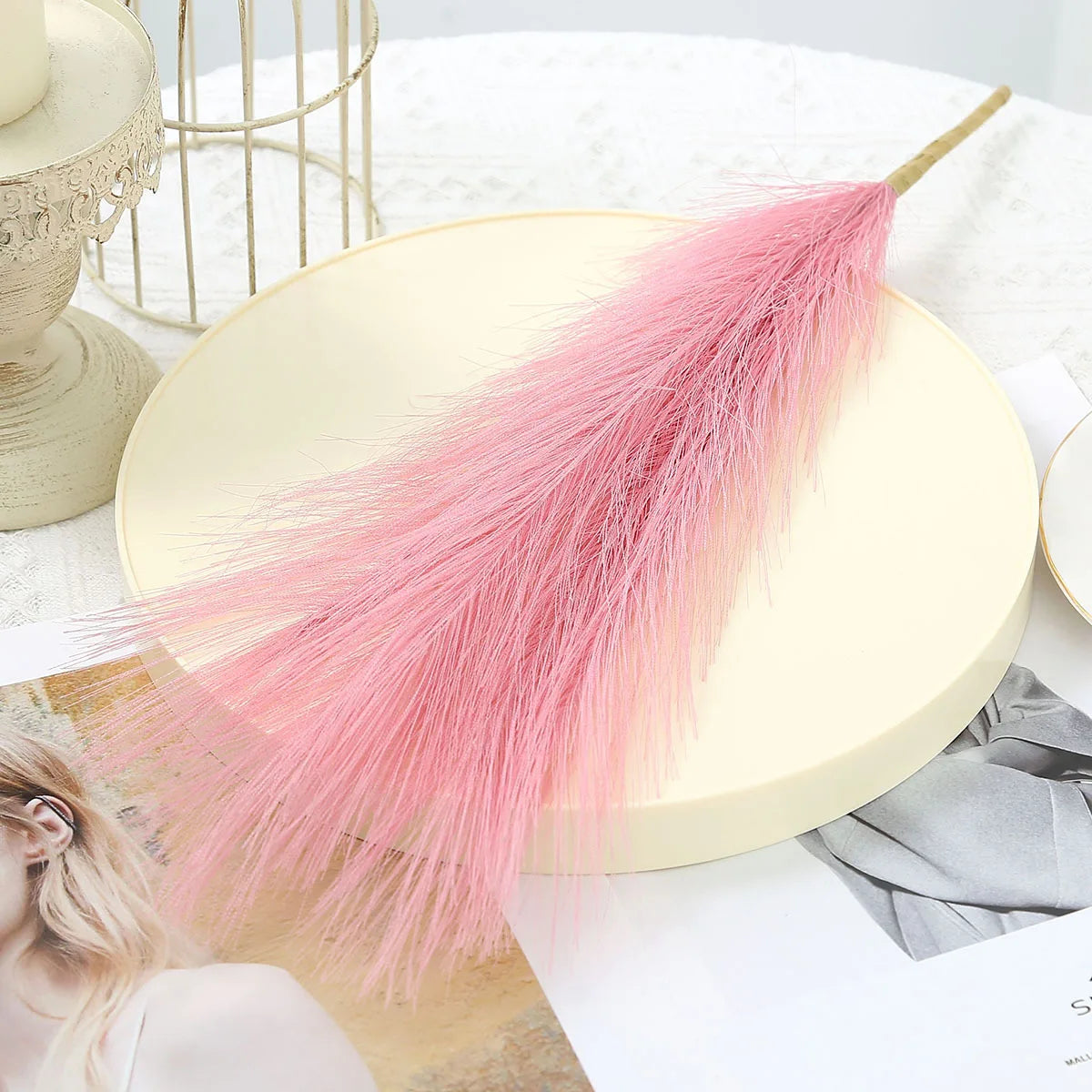 A pink artificial pampas grass bouquet displayed on a light yellow plate with natural decor elements.
