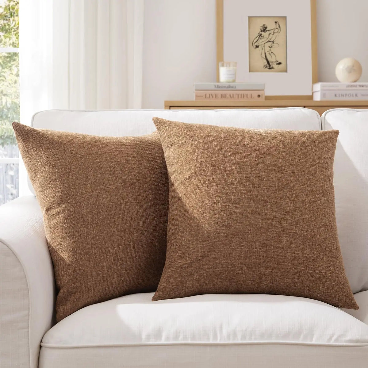 Two brown linen pillows resting on a white sofa with a framed picture in the background.