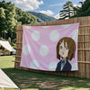 A pink tapestry featuring an anime character from K-On! with large white polka dots, displayed outdoors against a bamboo fenc