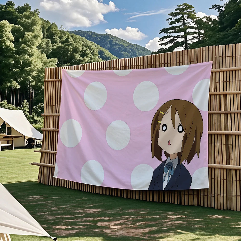 A pink tapestry featuring an anime character from K-On! with large white polka dots, displayed outdoors against a bamboo fenc