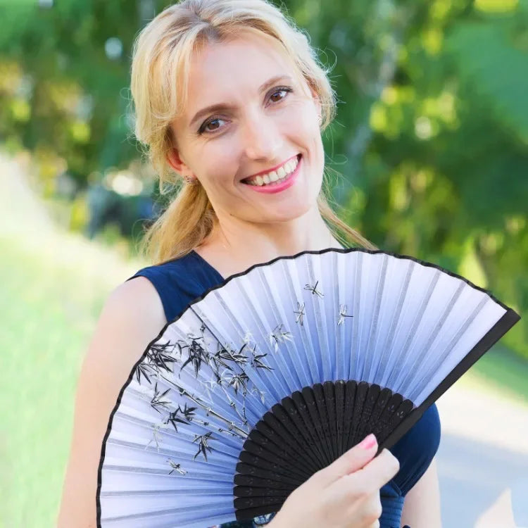 A woman holds a vintage bamboo folding hand fan with delicate silver accents against a blurred green background.