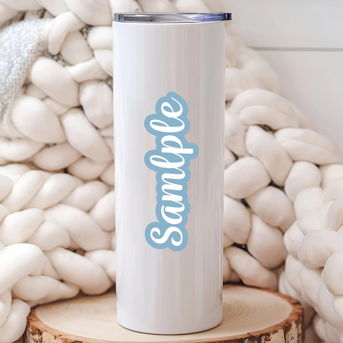 A white tumbler with light blue text that reads Sample is displayed against a background of cream-colored yarn.