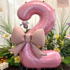 A large pink foil balloon shaped like the number 2 with a matte bow, set against green foliage and flowers.