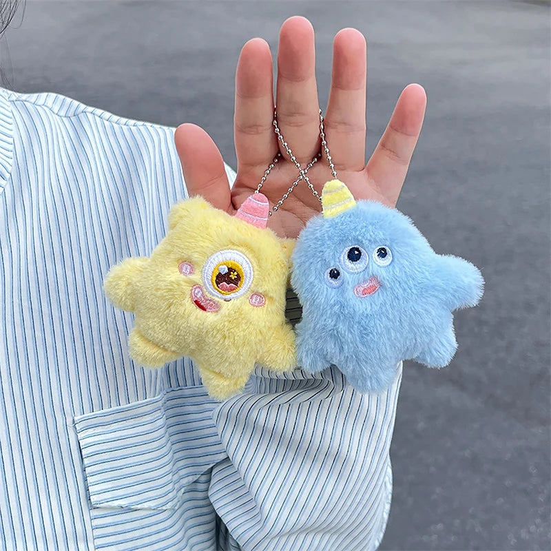Two fuzzy plush monster keychains, one yellow and one blue, are held by a person against a striped shirt.