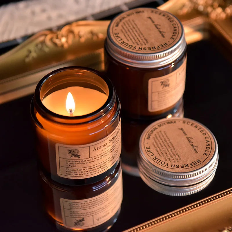 A hand-poured soy aromatherapy candle in a glass jar with a lit wick, set against a gold-edged box.