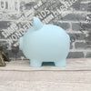 A light blue piggy bank shaped like a pig sitting on a wooden surface against a brick wall background.