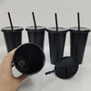 A hand holds a black reusable tumbler with a lid and straw, next to several other similar tumblers.