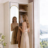 A full-length arched wall mirror with an aluminum frame reflecting a woman and displaying clothing storage.