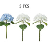 Three artificial hydrangea flower stems, two blue and one white, on green leaves.