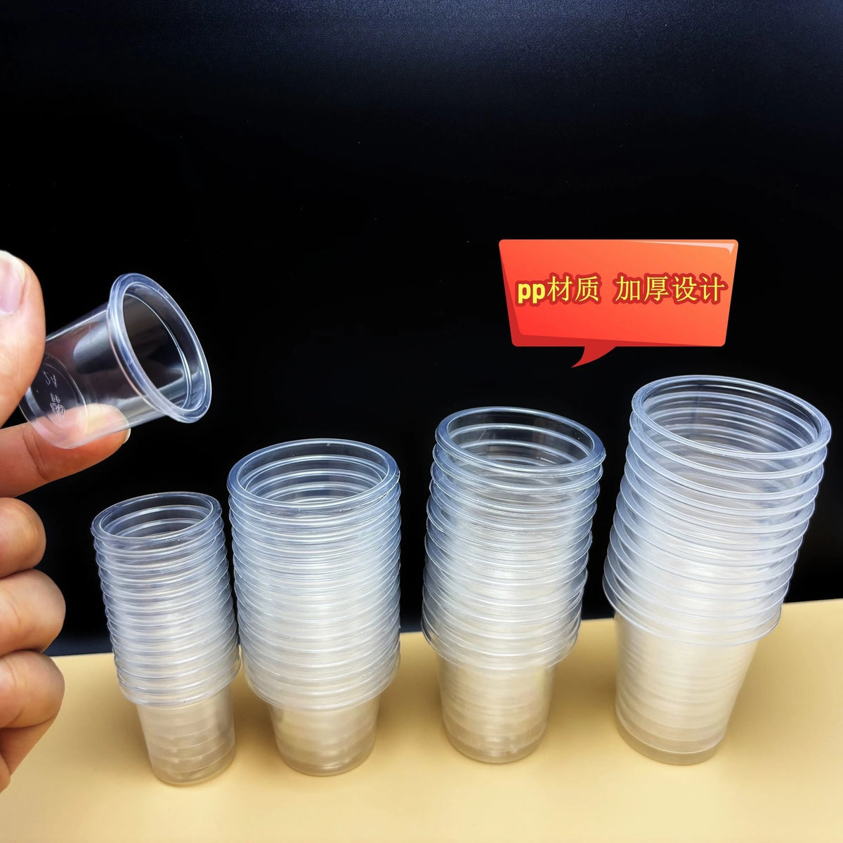 A hand holds a stack of clear, disposable mini tasting cups on a light surface against a black background.