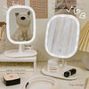 A white rechargeable LED vanity mirror with touch controls is displayed next to a cat figurine and skincare products.