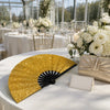 Gold and black foldable hand fans displayed on a white tablecloth at an outdoor event setting.
