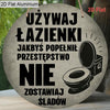 A round, dark gray aluminum sign with black text in Polish that reads: UŻYWAJ ŁAZIENKI JAKBYŚ POPEŁNIŁ PRZESTĘPSTWO NIE ZOSTA