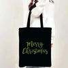 A black tote bag with green text that reads Merry Christmas, held by a person wearing a white sweater.