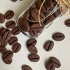 A glass jar filled with dark brown coffee bean cookies and twine, with loose cookies scattered on a white surface.