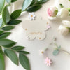 A white wooden monthly milestone card featuring a cloud shape with a pink rose and small flowers, surrounded by green eucalyp