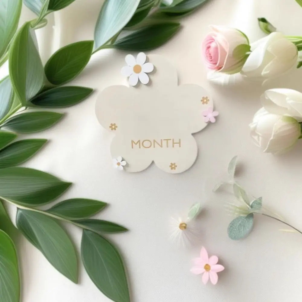 A white wooden monthly milestone card featuring a cloud shape with a pink rose and small flowers, surrounded by green eucalyp