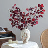 A vase holds a branch of autumn beech simulation flowers with deep red and purple leaves on a white table.
