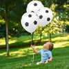 A baby is looking up at a cluster of white oval latex football party balloons floating in a sunny green park.