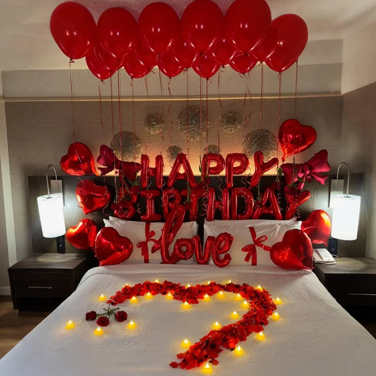 A bed decorated with red foil birthday balloons and a heart-shaped flower arrangement on the white bedding.