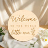 A round wooden welcome sign with the text Welcome to the world little one on a white cloth.
