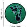 A round, dark green embroidered patch featuring a black silhouette of a Bigfoot foot.