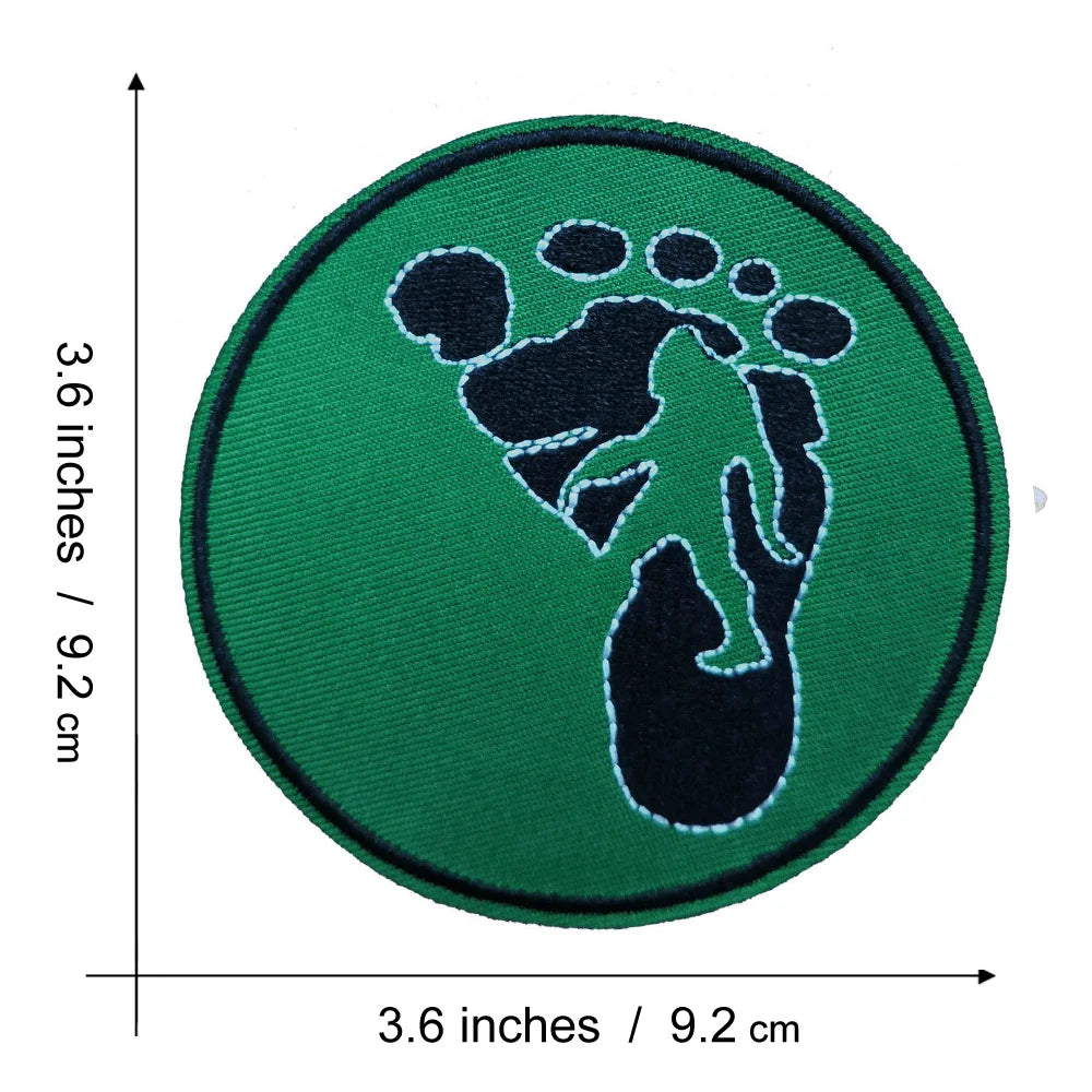 A round, dark green embroidered patch featuring a black silhouette of a Bigfoot foot.