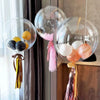 A collection of clear inflatable balloons in various shades of pink, gold, and black are displayed against a dark curtain.
