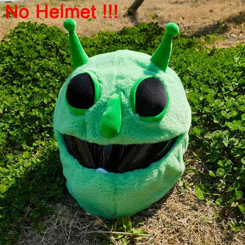 A bright green cartoon plush helmet cover with large black eyes and horns lying on dry grass.