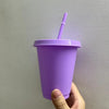 A hand holds a purple BPA-free plastic cold cup with a lid and straw against a light gray wall.