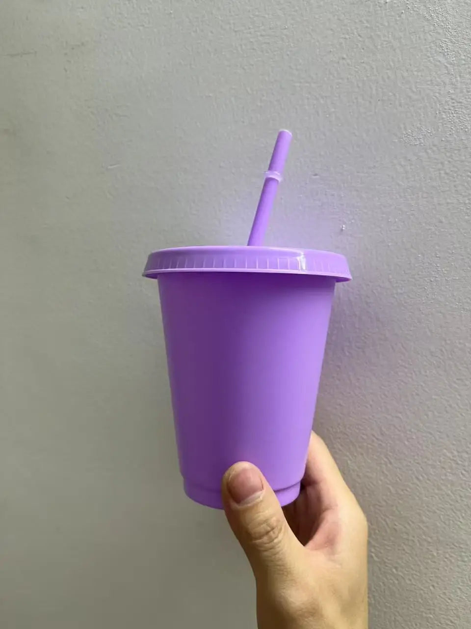 A hand holds a purple BPA-free plastic cold cup with a lid and straw against a light gray wall.
